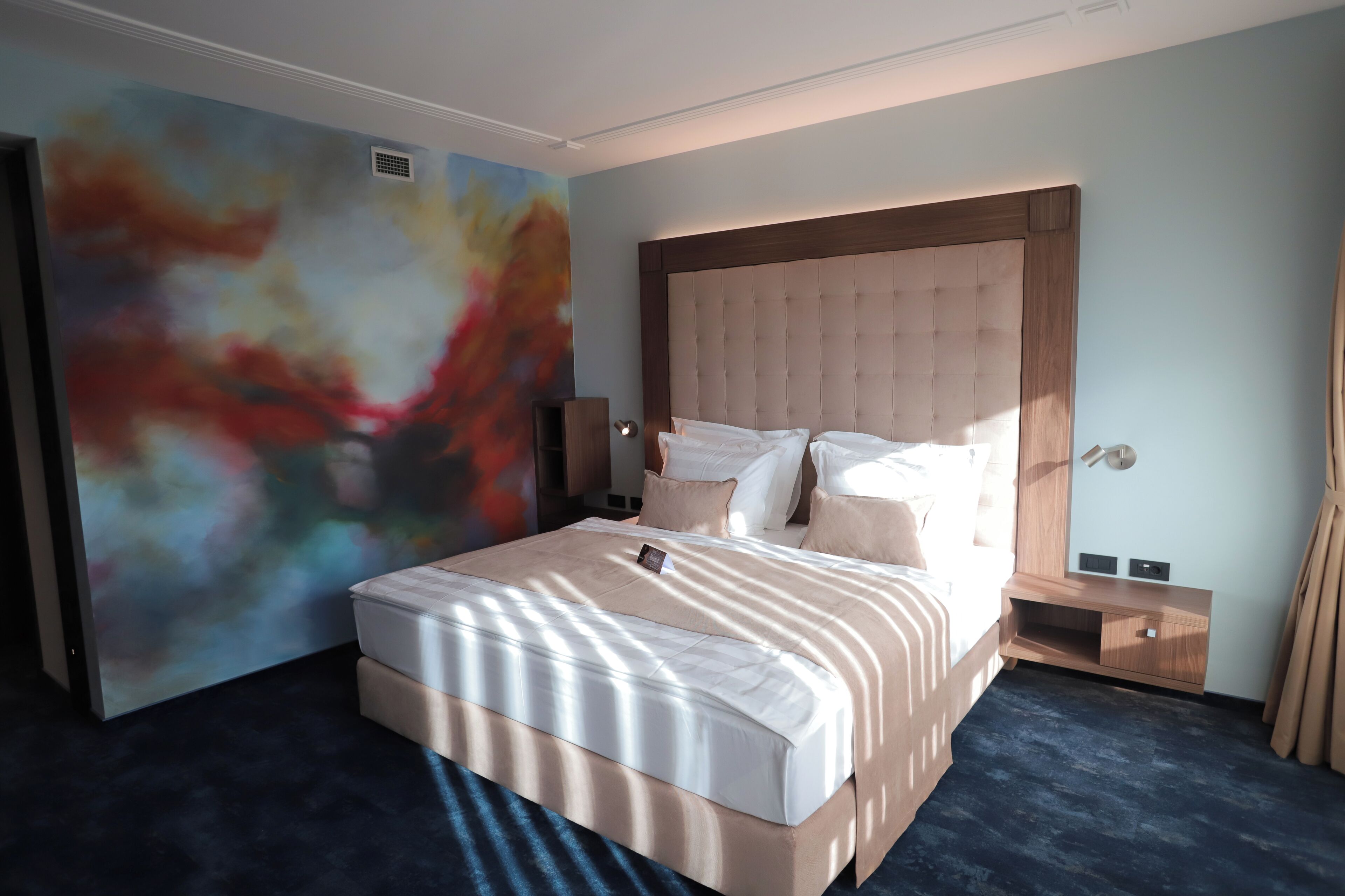 A modern hotel room with a large bed, a headboard, a bedside table, and a wall with abstract art.