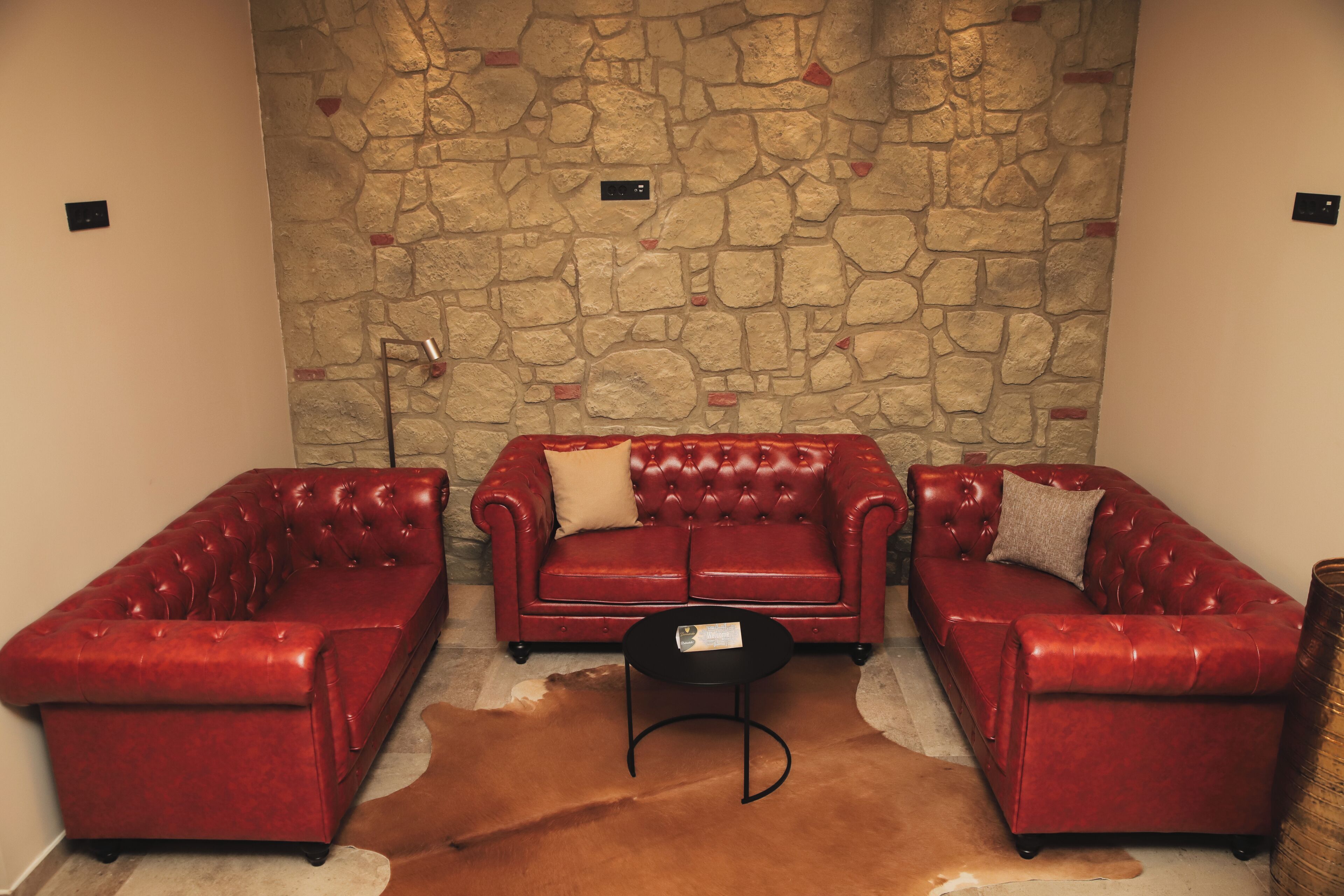 A room with a stone wall, three red leather sofas, a black coffee table, and a brown leather rug.