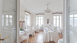 Une salle à manger élégante, avec des tables et des chaises blanches, un lustre et de grandes fenêtres.