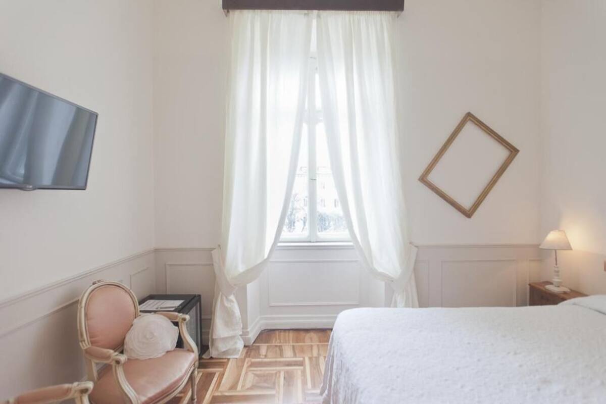 A hotel room with a bed, a chair, a TV mounted on the wall, a window with curtains, and a bedside table with a lamp.