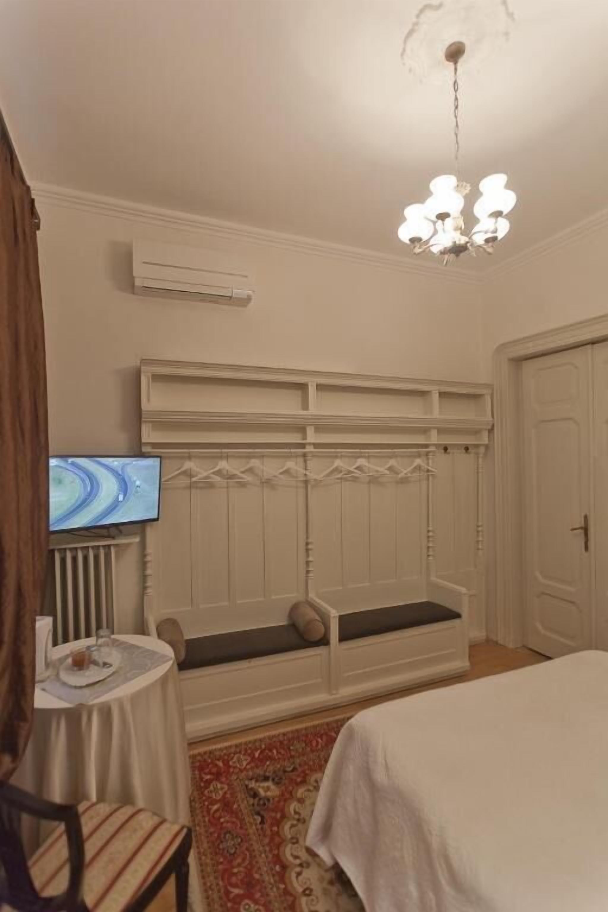 A hotel room with a bed, a television on a stand, a small table with a chair, a bench with storage, and a ceiling light fixture.