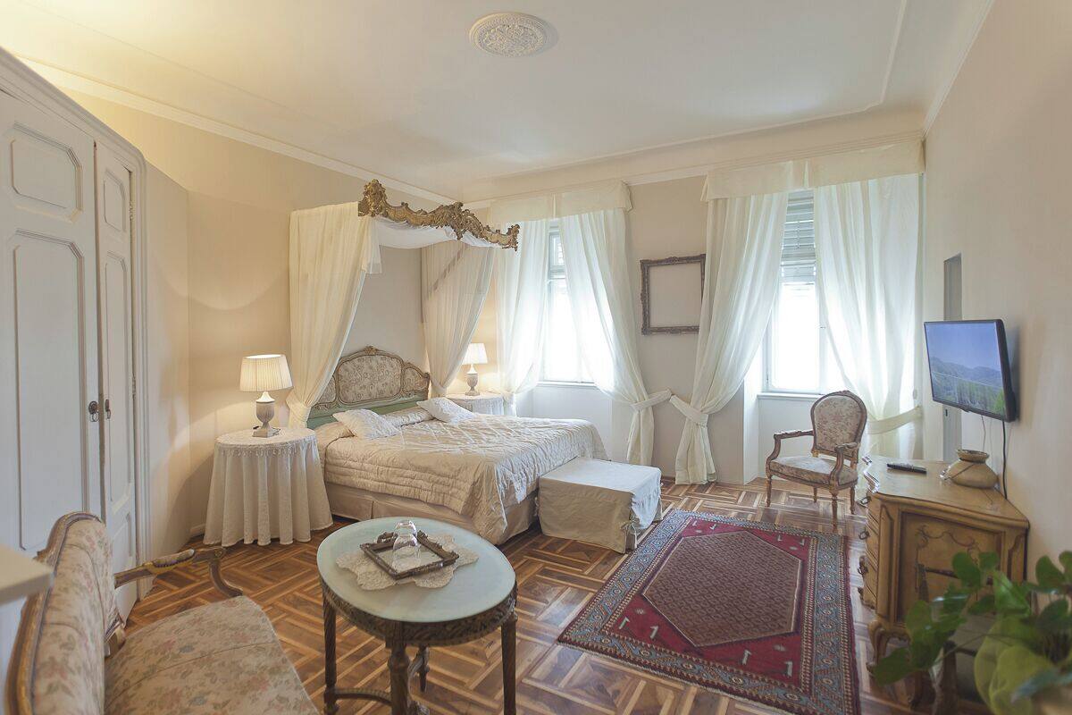 A bedroom with a four-poster bed, a sitting area with a table and chair, a television, and a large window with curtains.