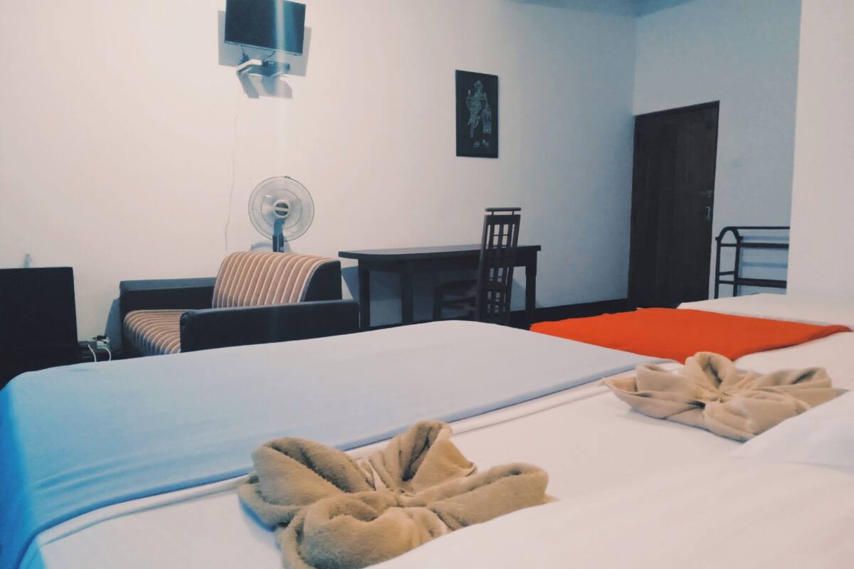 A hotel room with two beds, a desk, a chair, a TV, and a fan.
