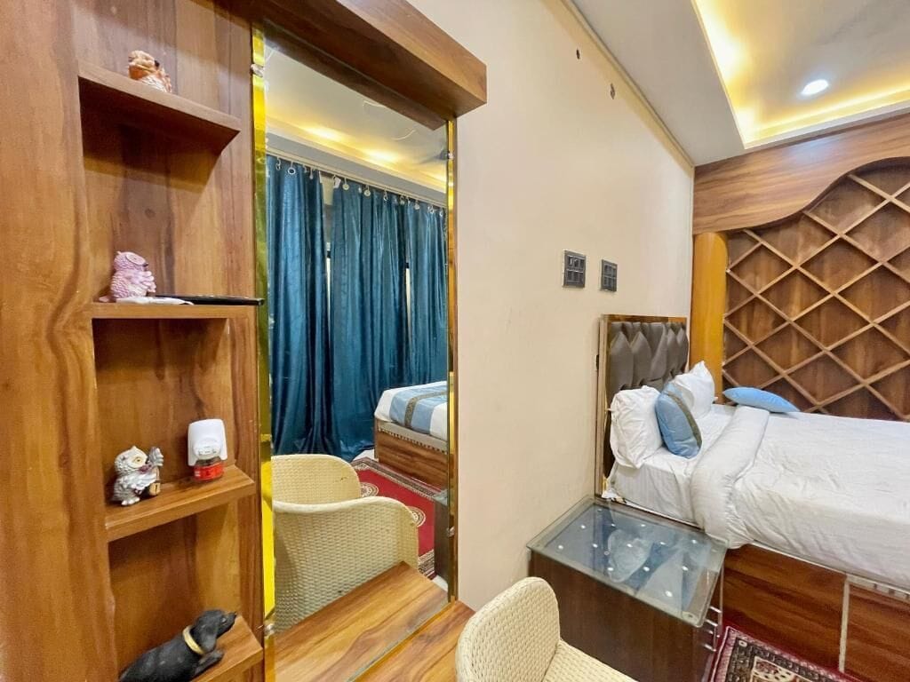 A hotel room with a wooden wardrobe, a bed with a headboard, a small table, a chair, and a mirror.