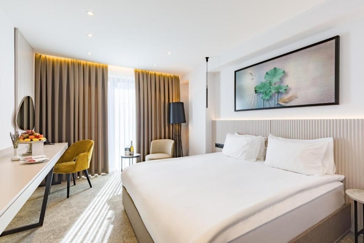 A hotel room with a large bed, a desk with a chair, a small table, and a wall-mounted artwork.
