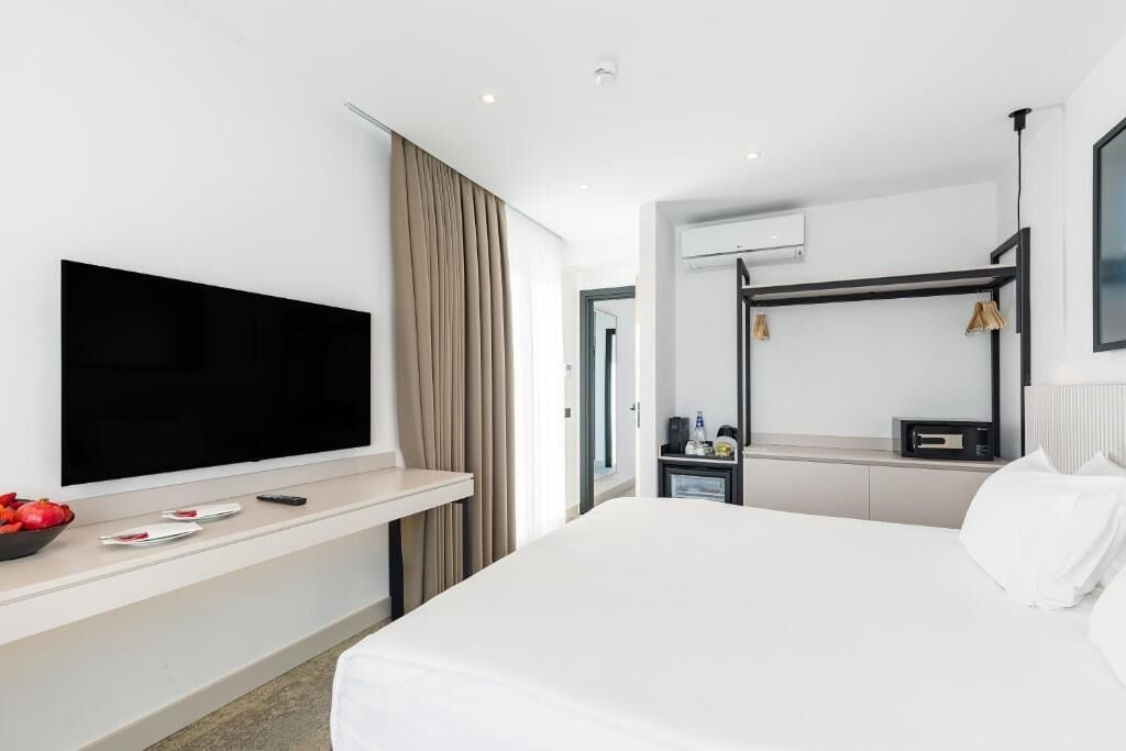 A modern hotel room with a large bed, a flat-screen TV mounted on a wall, a desk with a chair, and a bunk bed.