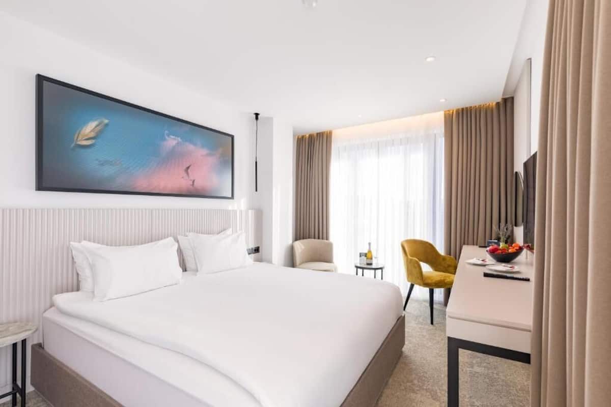 A modern hotel room with a large bed, a desk with a chair, a painting on the wall, and a window with curtains.