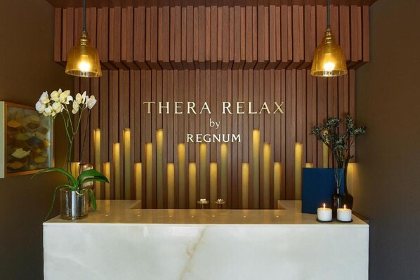 A reception desk with a wooden backdrop, two hanging lamps, and a sign that reads "THERA RELAX by REGNUM".