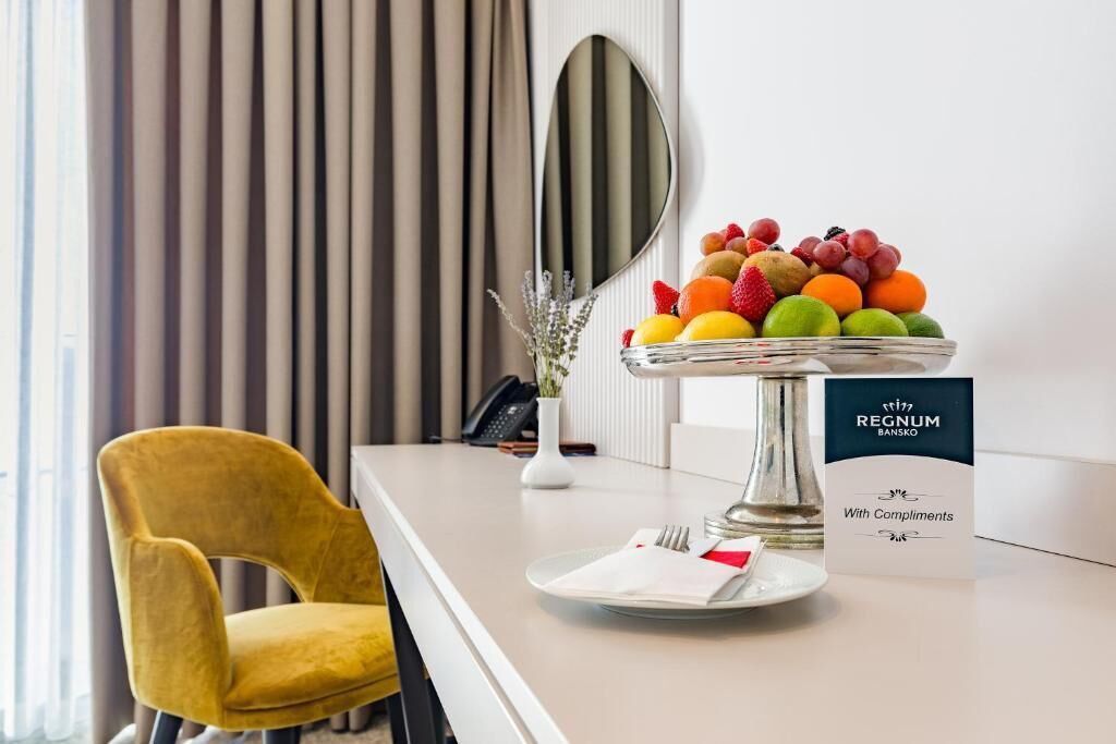 A hotel reception area with a fruit bowl, a chair, a mirror, and a sign reading "REGNUM BANSKO With Compliments".