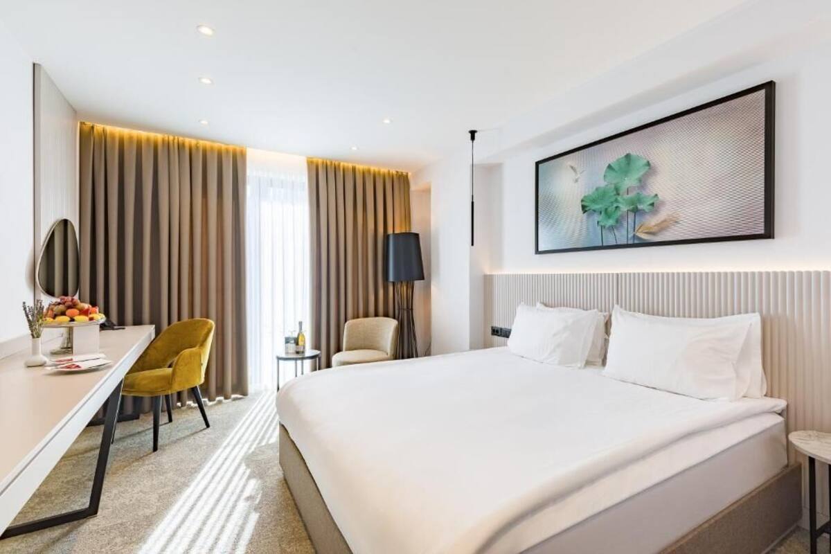 A hotel room with a large bed, a desk with a chair, a small table, and a wall-mounted artwork.