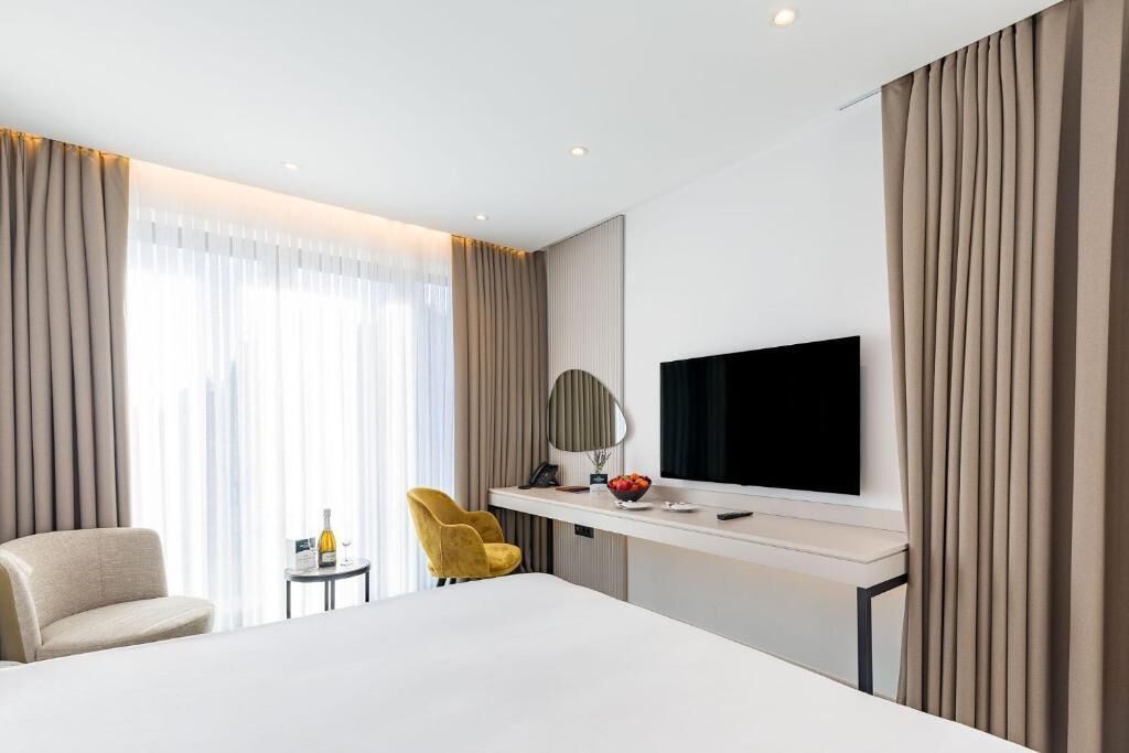 Una habitación de hotel con una cama grande, un escritorio con televisor de pantalla plana, una silla y una mesita con una botella y fruta.