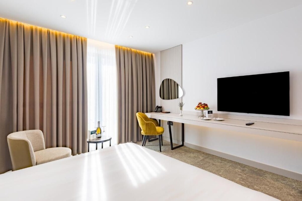 A hotel room with a large bed, a desk with a flat-screen TV, a chair, and a small table with a bottle and fruit.