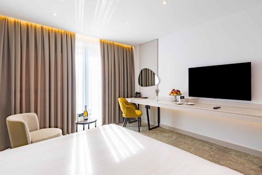 Habitación de hotel con una cama grande, un escritorio con televisor de pantalla plana, una silla y una mesita con una botella y fruta.