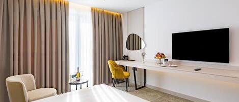 A hotel room with a large bed, a desk with a flat-screen TV, a chair, and a small table with a bottle and fruit.