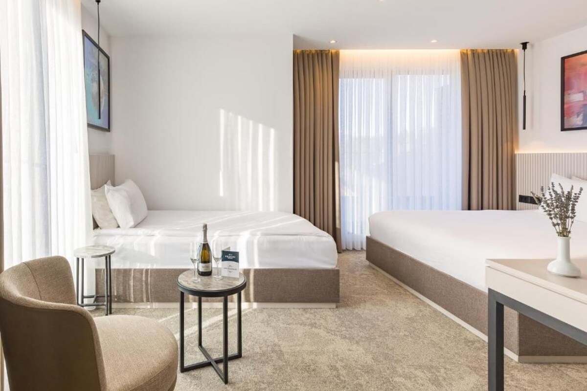 A hotel room with a bed, a small table with a bottle and glass, a chair, and a bedside table with a vase.