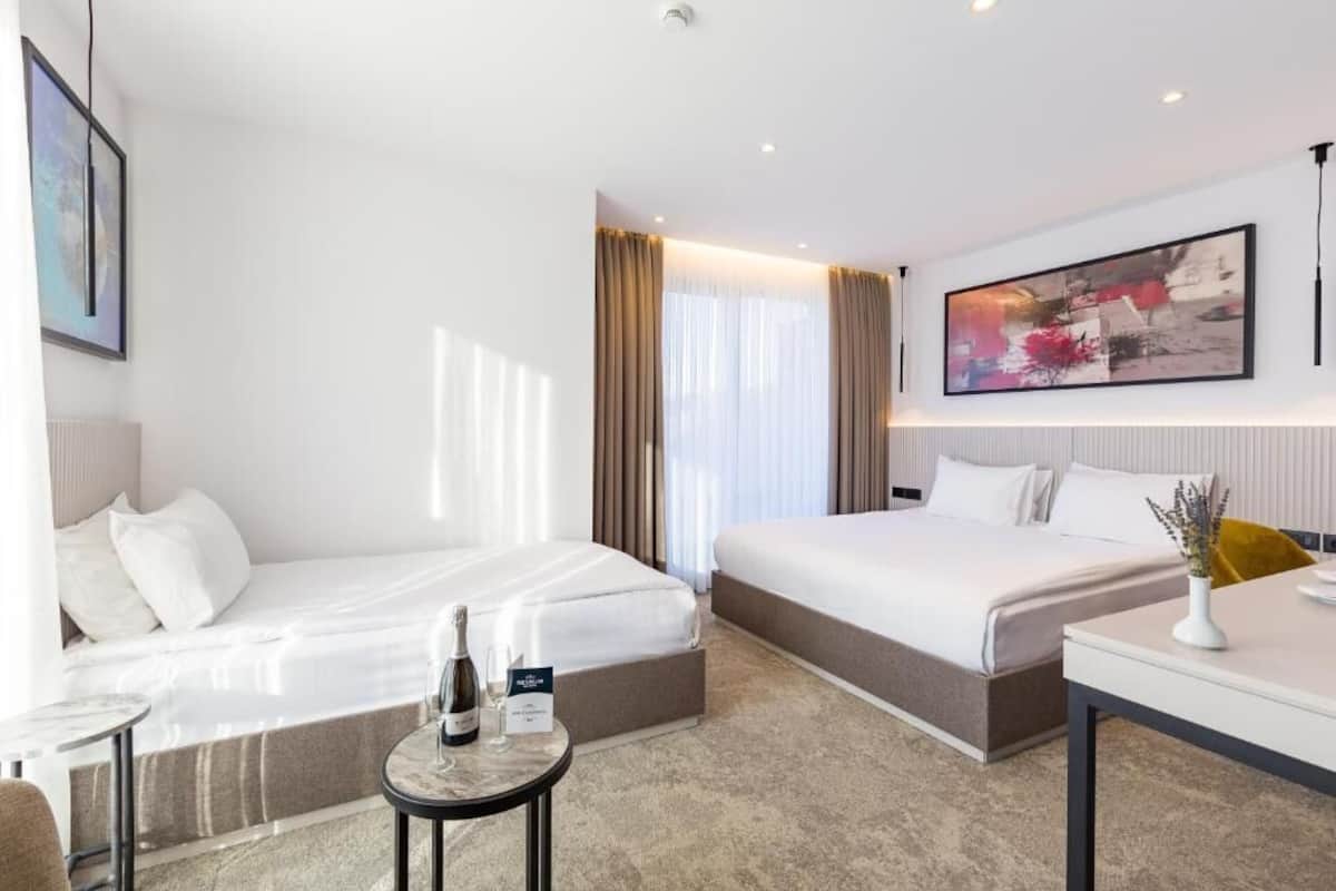 A hotel room with two beds, a small table with a bottle and glasses, and a painting on the wall.