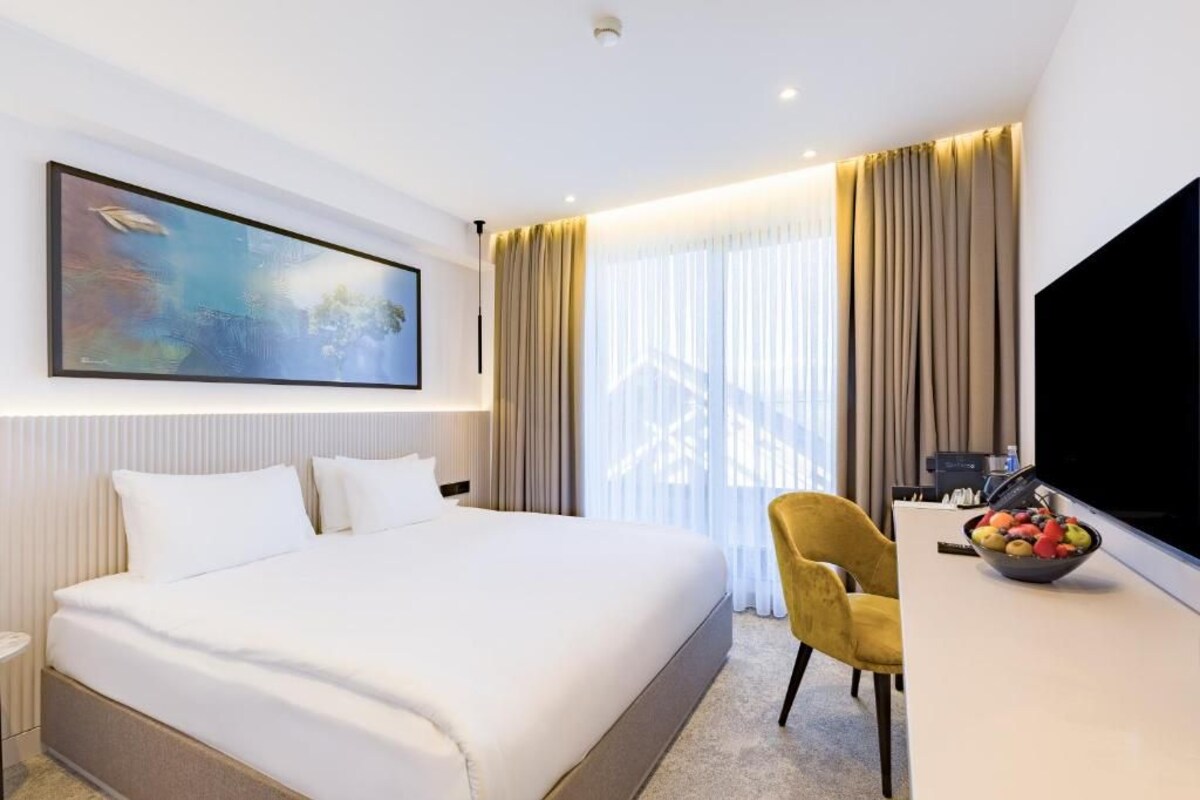 A modern hotel room with a large bed, a flat-screen TV, a dining table with fruit, and a painting on the wall.