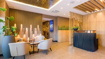 A modern reception area with a wooden reception desk, a seating area with two chairs and a small table, and a large potted plant.
