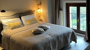 Une chambre à coucher avec un grand lit, une table de chevet, une lampe et une vue sur la verdure par la fenêtre.