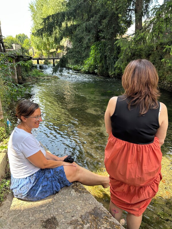 Two individuals seated by a calm river with a bridge in the background.