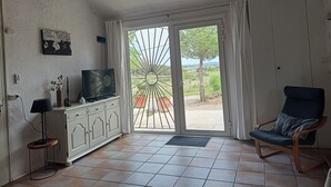 A room with a television on a cabinet, a chair, and a view through a glass door to an outdoor area with trees.