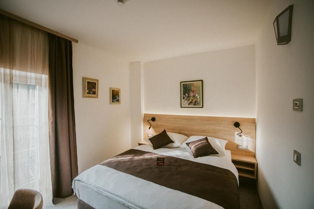A hotel room with a large bed, two bedside tables, a nightstand, a window with curtains, and two wall-mounted lamps.