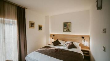 A hotel room with a large bed, two bedside tables, a nightstand, a window with curtains, and two wall-mounted lamps.