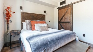 Ein Schlafzimmer mit einem Holzkopfende, einem großen Bett mit grauer Decke, einem Nachttisch mit einer Vase getrockneter Blumen und einer massiven Schiebetür im Scheunenstil.