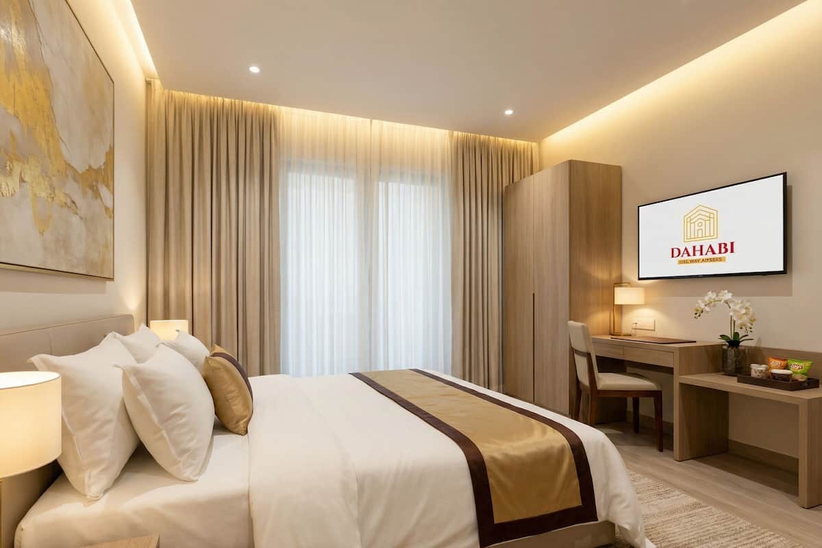Una habitación de hotel con cama, escritorio, silla y televisor mostrando el logo de Dahabi.