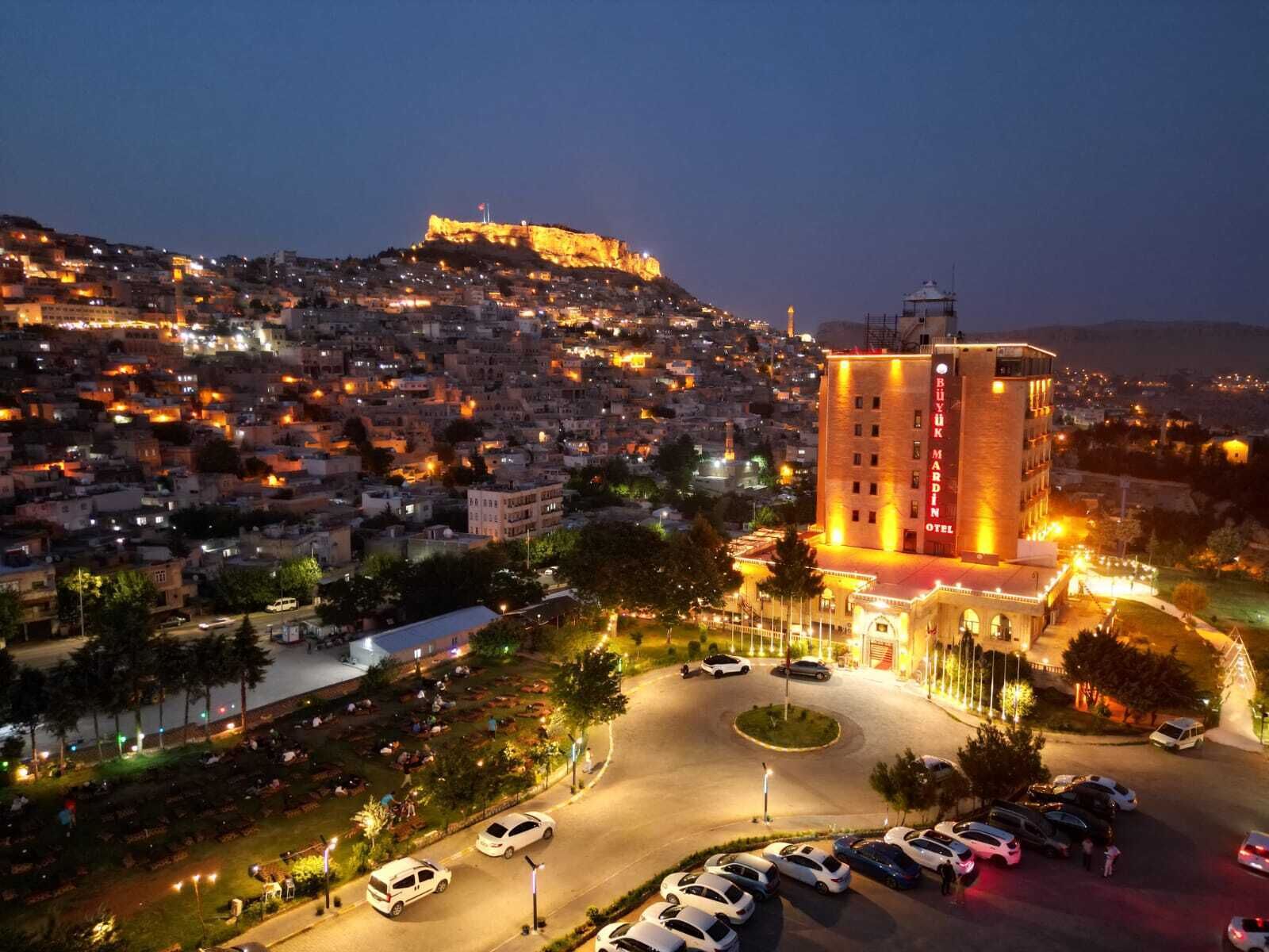 A hotel with a prominent tower, surrounded by a parking area and a view of a densely populated cityscape with a hill in the background.