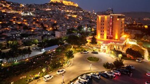 A hotel with a prominent tower, surrounded by a parking area and a view of a densely populated cityscape with a hill in the background. - Büyük Mardin Otel (Mardin)