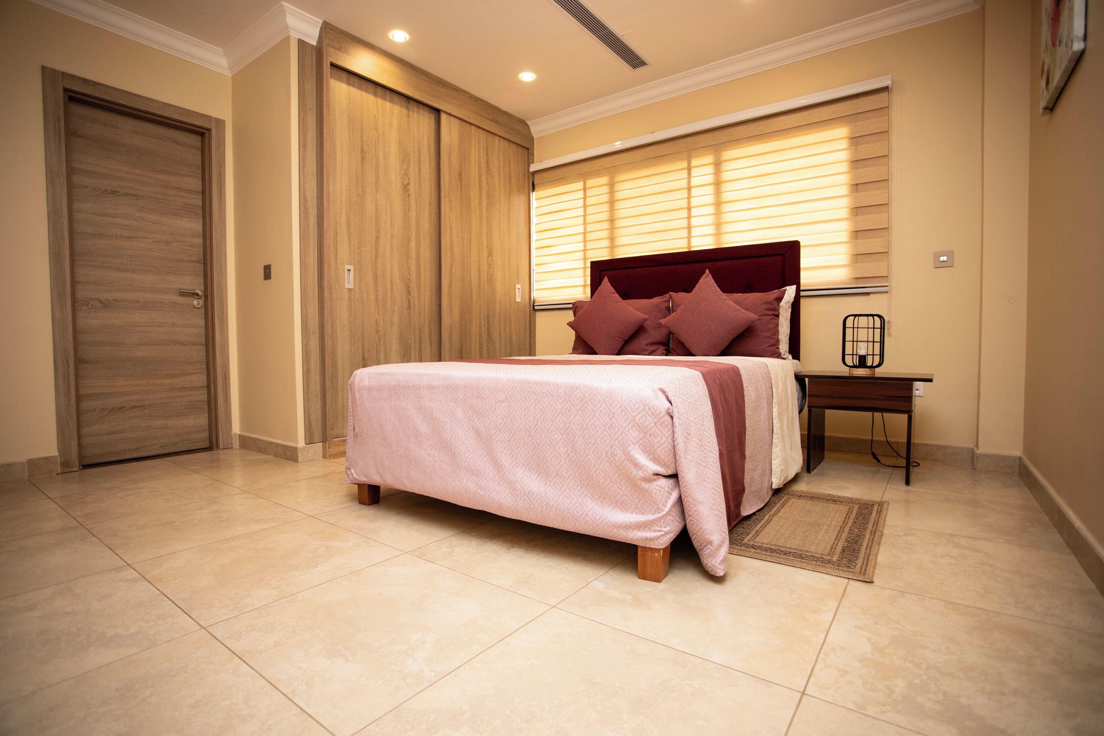 A bedroom with a bed, a nightstand, a wooden wardrobe, and a window with blinds.