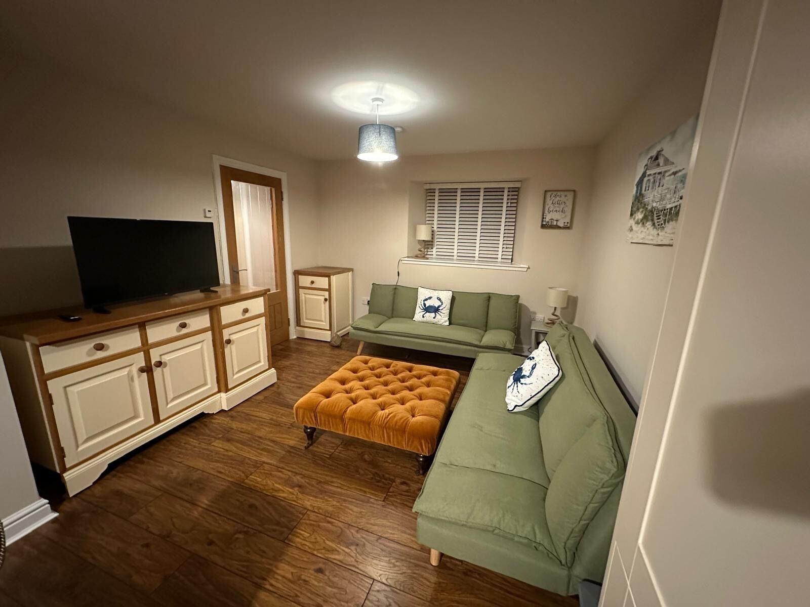 A compact living space with a television on a wooden cabinet, a green sofa with a crab-themed pillow, a tufted ottoman, and a small side table.