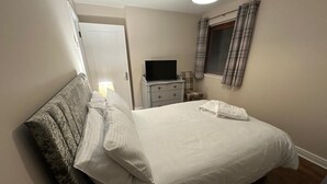 Een netjes opgemaakt bed met kussens, een nachtkastje met een lamp, een televisie en een raam met gordijnen.