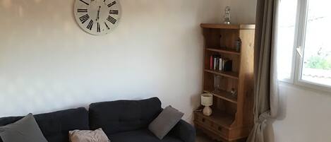 A living room with a dark-colored sofa, a wooden side table with a bowl and a plant, a wooden bookshelf, and a wall clock.