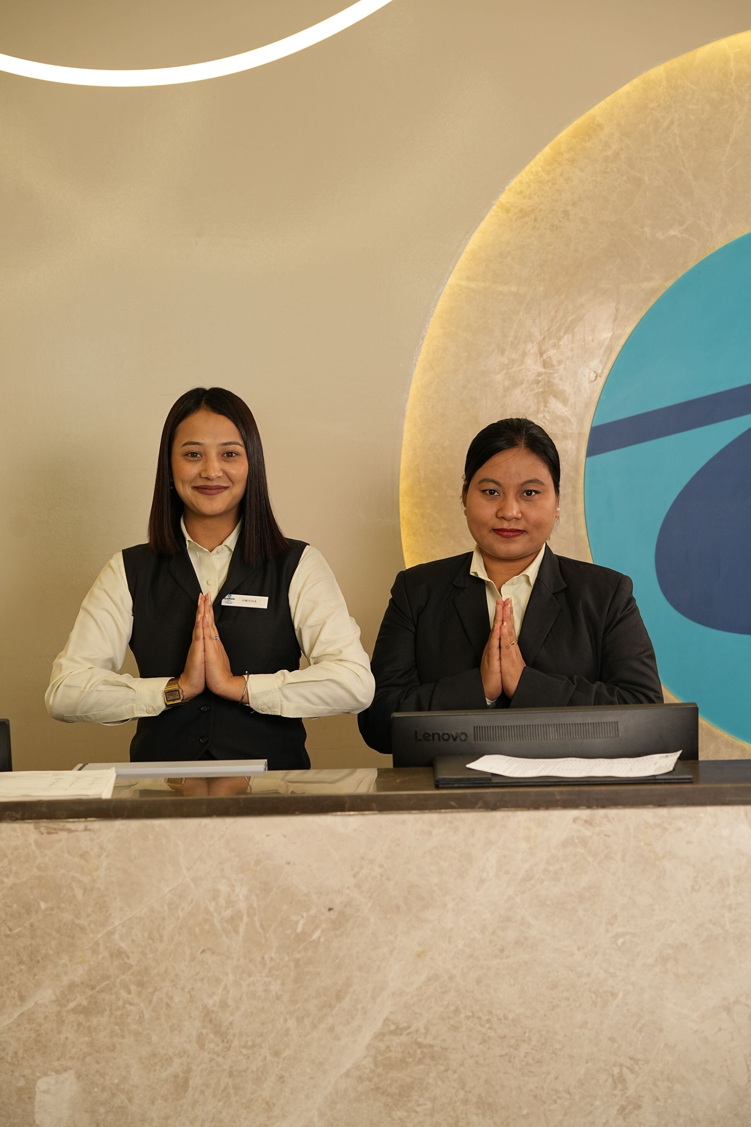 Two receptionists standing behind a reception desk with a laptop and a name tag.