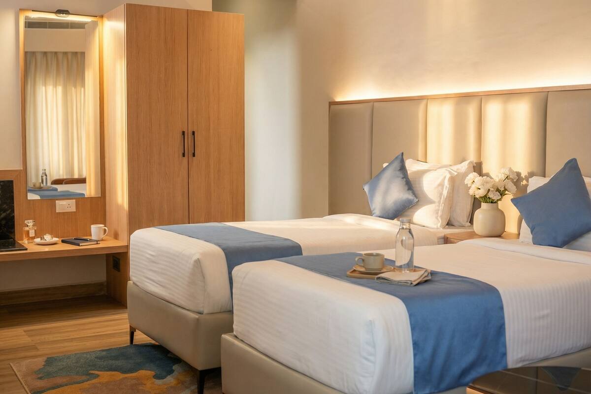 A hotel room with two beds, a wooden wardrobe, a desk with a chair, a mirror, and a nightstand with a vase of flowers.