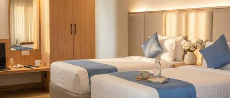 A hotel room with two beds, a wooden wardrobe, a desk with a chair, a mirror, and a nightstand with a vase of flowers.