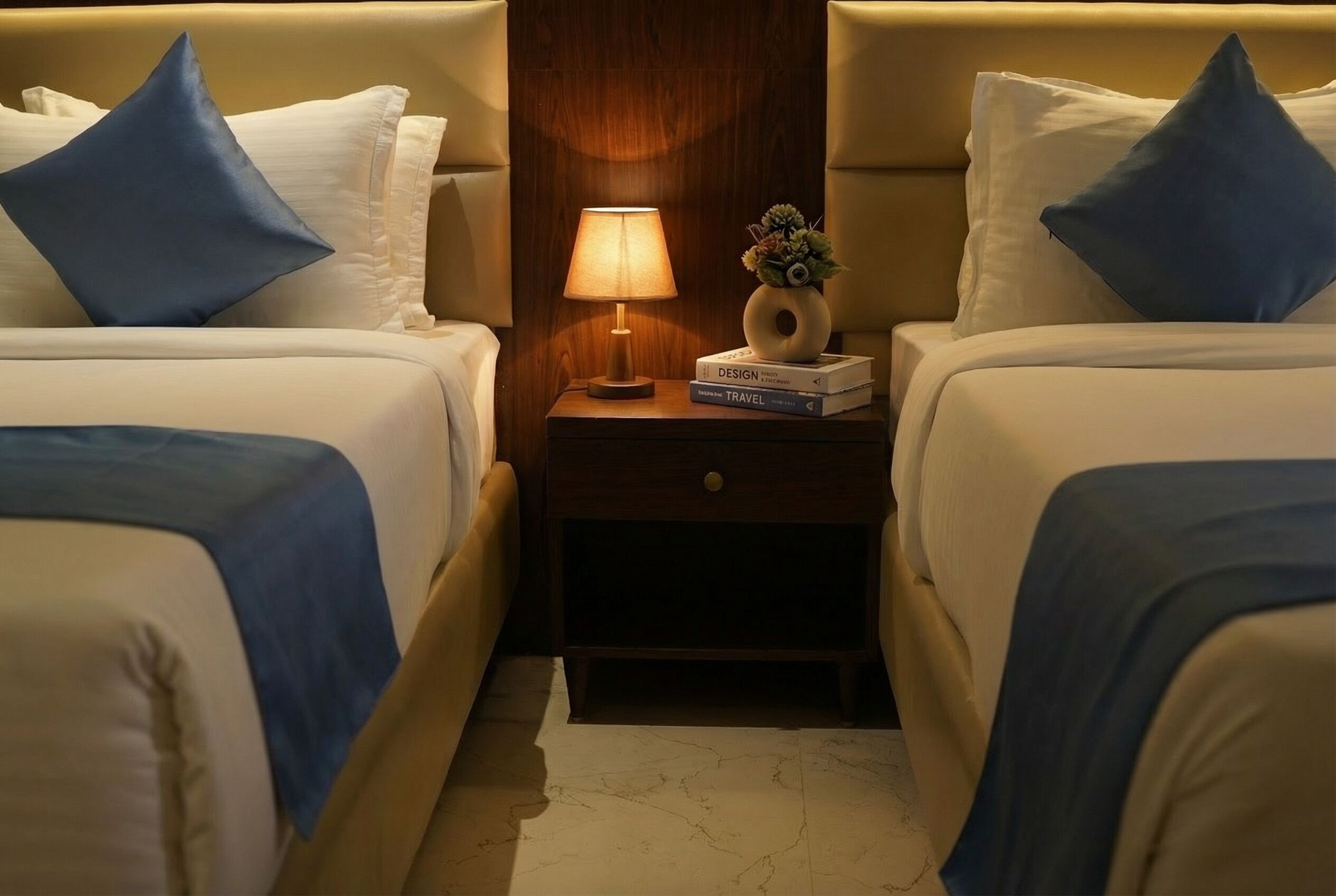 A hotel room with two beds, a nightstand with a lamp, and a small vase with flowers.