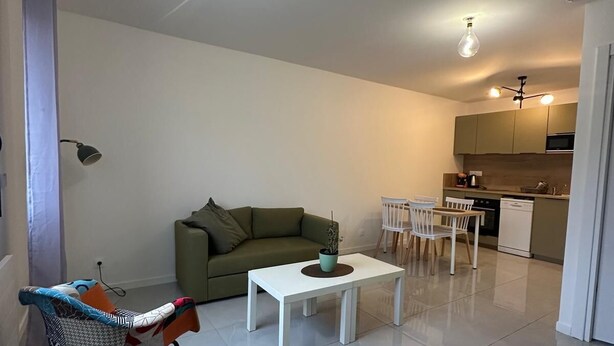 Ein modernes Wohnzimmer mit einem grünen Sofa, einem weißen Couchtisch und einem Schaukelstuhl. Es gibt eine Küchenzeile mit einem Esstisch und Stühlen.