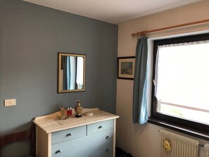Une pièce comprenant une commode, un miroir, une fenêtre avec des rideaux, un radiateur et un tableau accroché au mur.