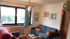 A living room with a blue patterned sofa, a red armchair, a small table with a laptop, and a window with curtains. - Suite Marco Nice Apartment in Cavalese With View (Cavalese)