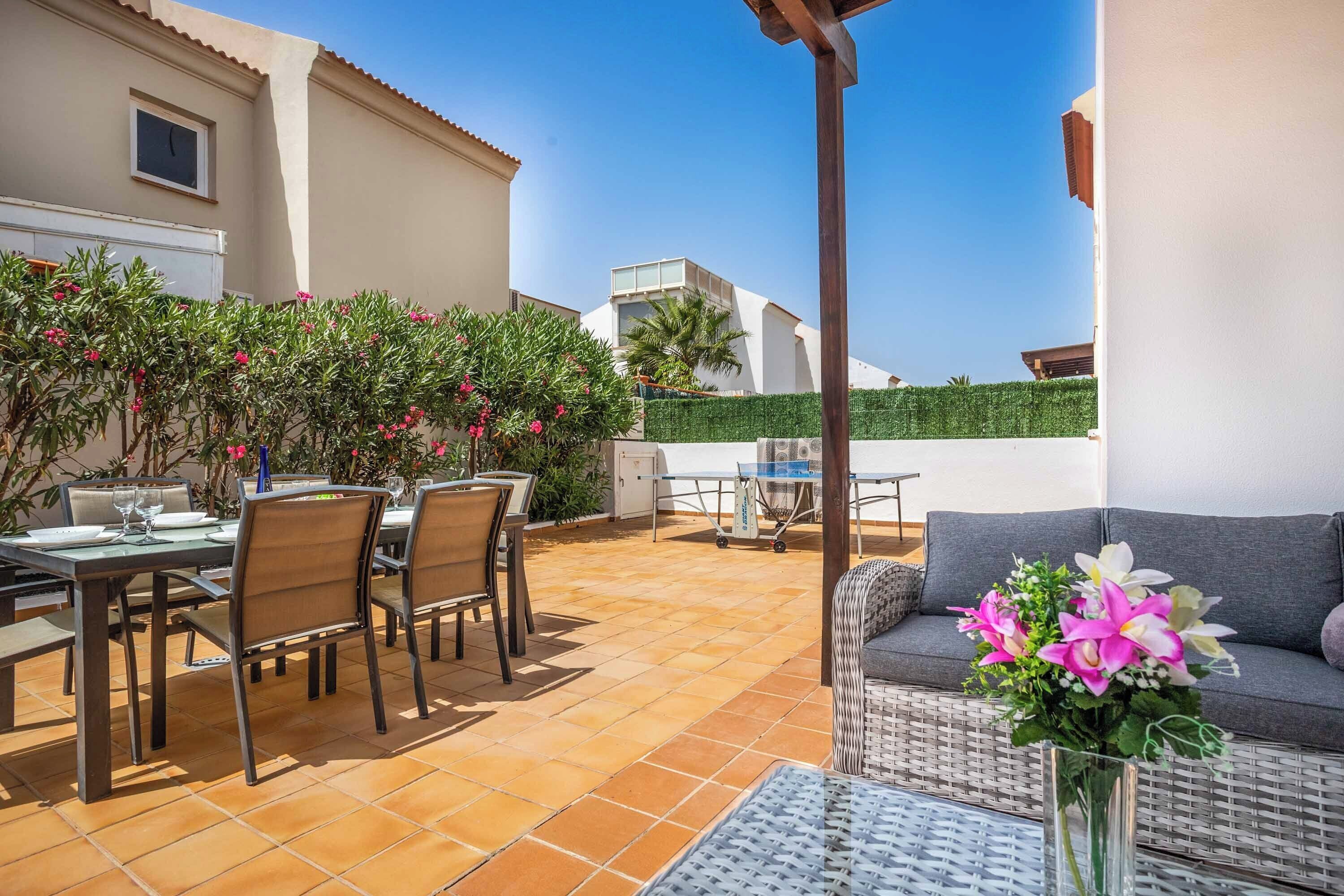 Eine Außenpatio mit Esstisch, Stühlen und einer Couch. Ein kleiner Tisch mit einem Blumenstrauß steht daneben.