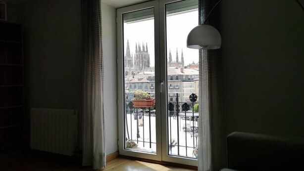 A room with a large sliding glass door leading to a balcony, a radiator, a lamp, and a view of a cityscape with a cathedral.