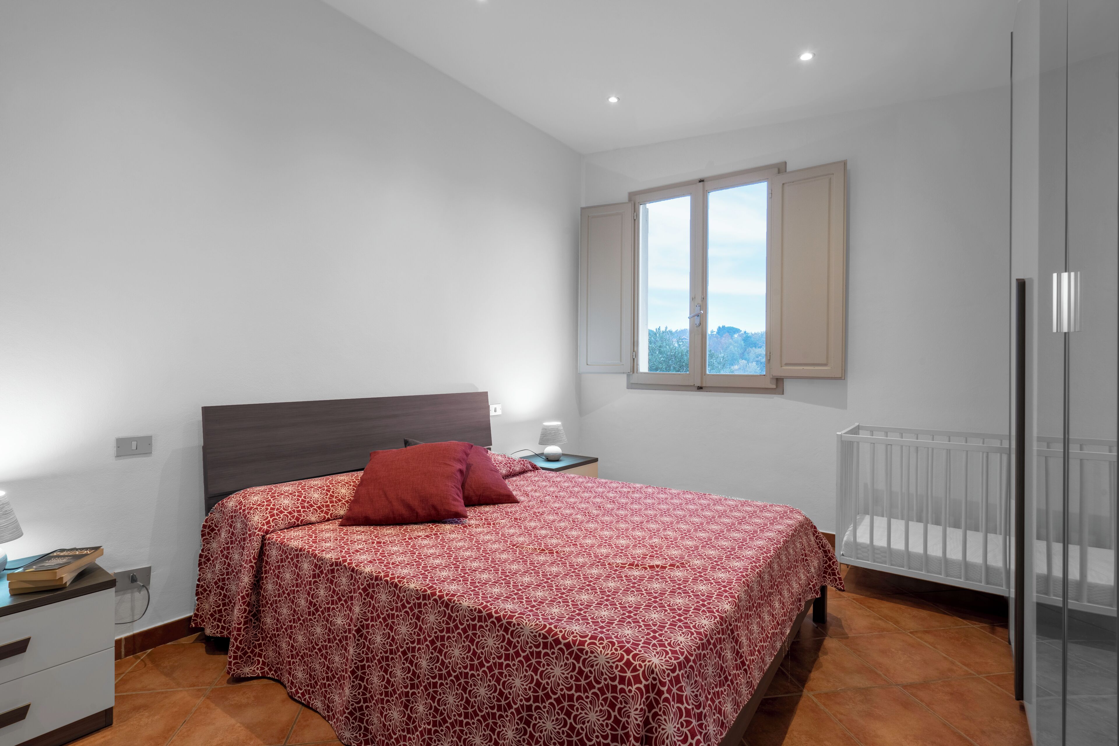 Ein Schlafzimmer mit einem Bett, einem Nachttisch, einer Lampe, einem Fenster mit Aussicht und einem Babybett.