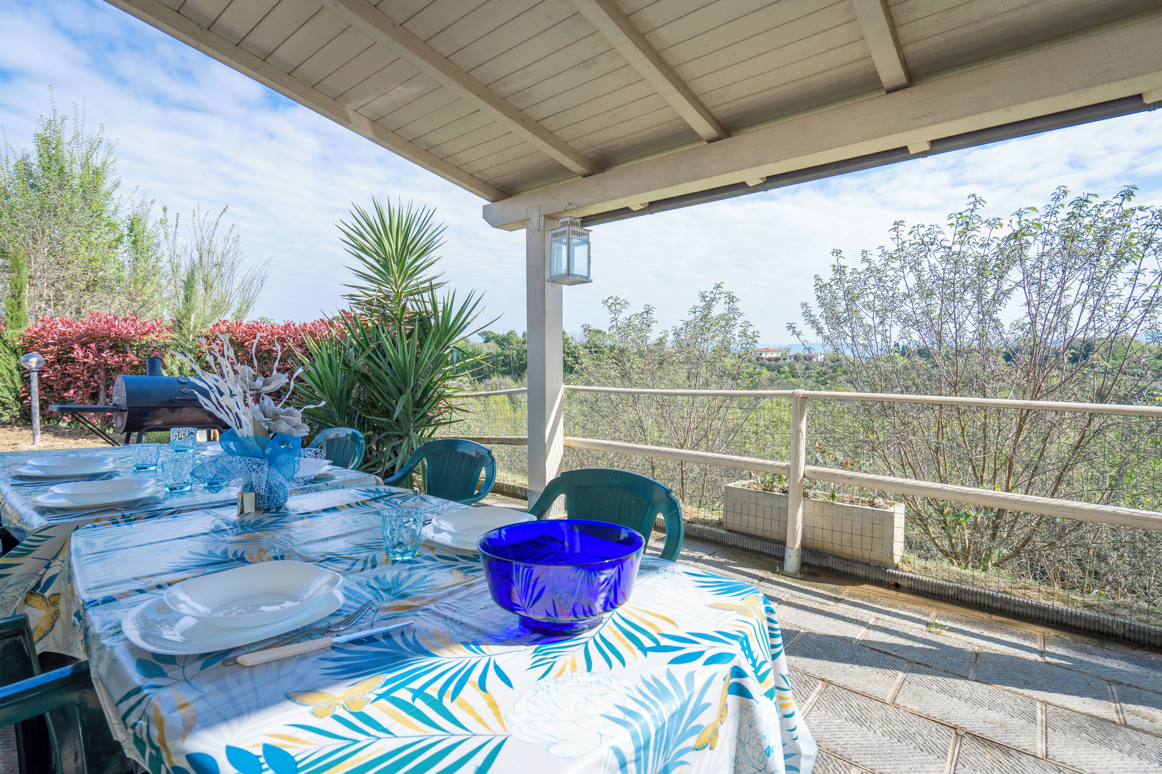 Un área de comedor al aire libre con un porche cubierto, una mesa con platos, vasos y un cuenco azul, rodeada de sillas y plantas en macetas.