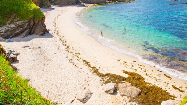 Een zandstrand met helder turquoise water, rotsachtige rotsformaties en schaarse begroeiing.