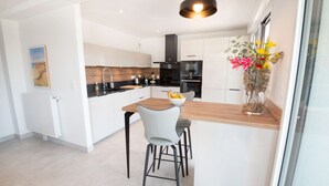 Een moderne keuken met witte kastjes, een houten aanrechtblad en een zwarte achterwand. Er is een eethoek met een tafel en twee stoelen. Op het aanrecht staat een vaas met bloemen.