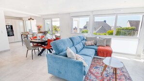 Een moderne woonkamer met een blauwe bank, een eethoek met rode stoelen en grote ramen met uitzicht op de omgeving.