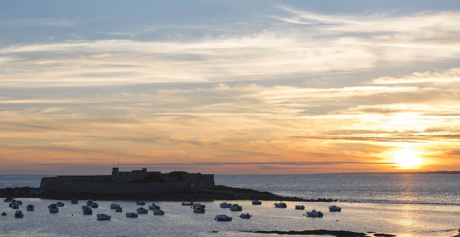 A coastal scene with a sunset, boats, and a fortified structure.